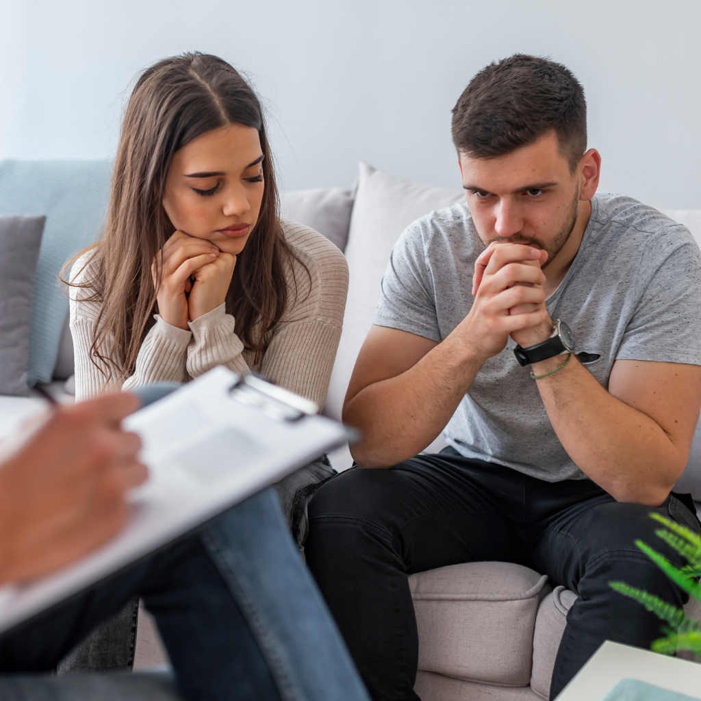 young adult man and woman sitting on a couch in a marriage counseling session. Marriage counselor can be seen, but is cropped out of the photo.